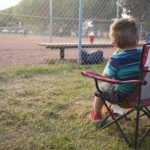 Little Boy sits in "Canada" child-sized lawn chair and watches Dad play baseball