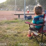 Little Boy sits in "Canada" child-sized lawn chair and watches Dad play baseball